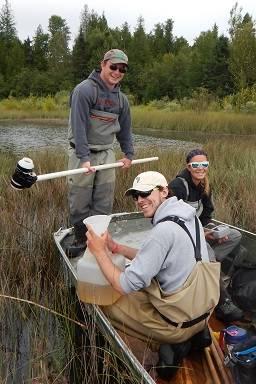 Three students display tools to measure water quality while sitting in a rowboat in a wetland.